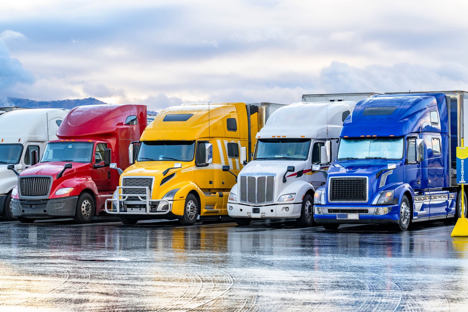 Different color and makes big rig semi trucks with semi trailers standing in row on the truck stop parking lot early morning with wet after rain surface